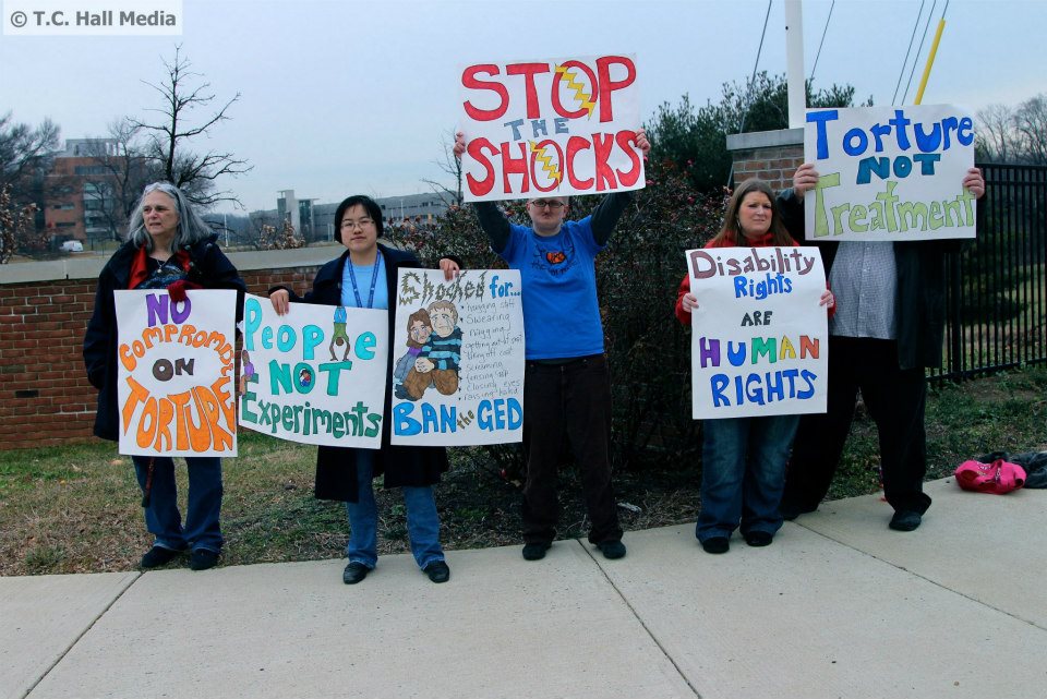 Photo: Five people (four white, one east asian) holding hand-made protest signs outside the Food and Drug Administration's White Oak Campus in Maryland. The signs say, "No Compromise on Torture," "People Not Experiments," "Shocked for... hugging staff, swearing, nagging, getting out of seat, taking off coat, screaming, tensing up, closing eyes, raising hand. BAN the GED.," "Stop the Shocks," "Disability Rights are Human Rights," and "Torture Not Treatment." Left to right: Diane Engster, Lydia X. Z. Brown, Shain M. Neumeier, Kathleen Nicole O'Neal, and Patrick T. Ayers. Photo by Taylor C. Hall. January 2013..